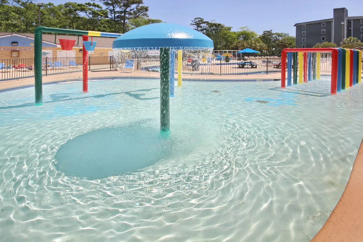 An exterior shot of a splash pad area with colorful water features and clear blue water, where children are seen playing and families are gathered nearby. The mood is joyful and sun-soaked, ideal for younger children.