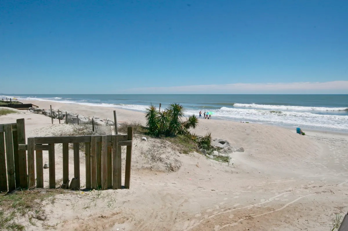 This image captures a peaceful stretch of beach with gentle ocean waves, a wooden fence in the foreground, palm-like plants, and a few people enjoying the shoreline in the distance. The scene is tranquil, offering a sense of openness and connection to nature.
