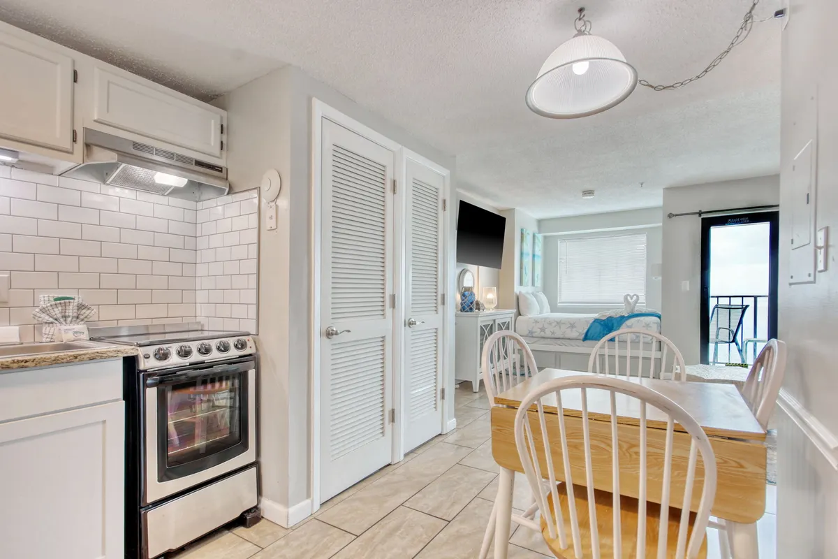 A view of the dining table set near a kitchen stove, with a plush couch and a balcony door in the background. The space is open and flows naturally from the kitchen to the living area, encouraging togetherness and easy access to outdoor views.