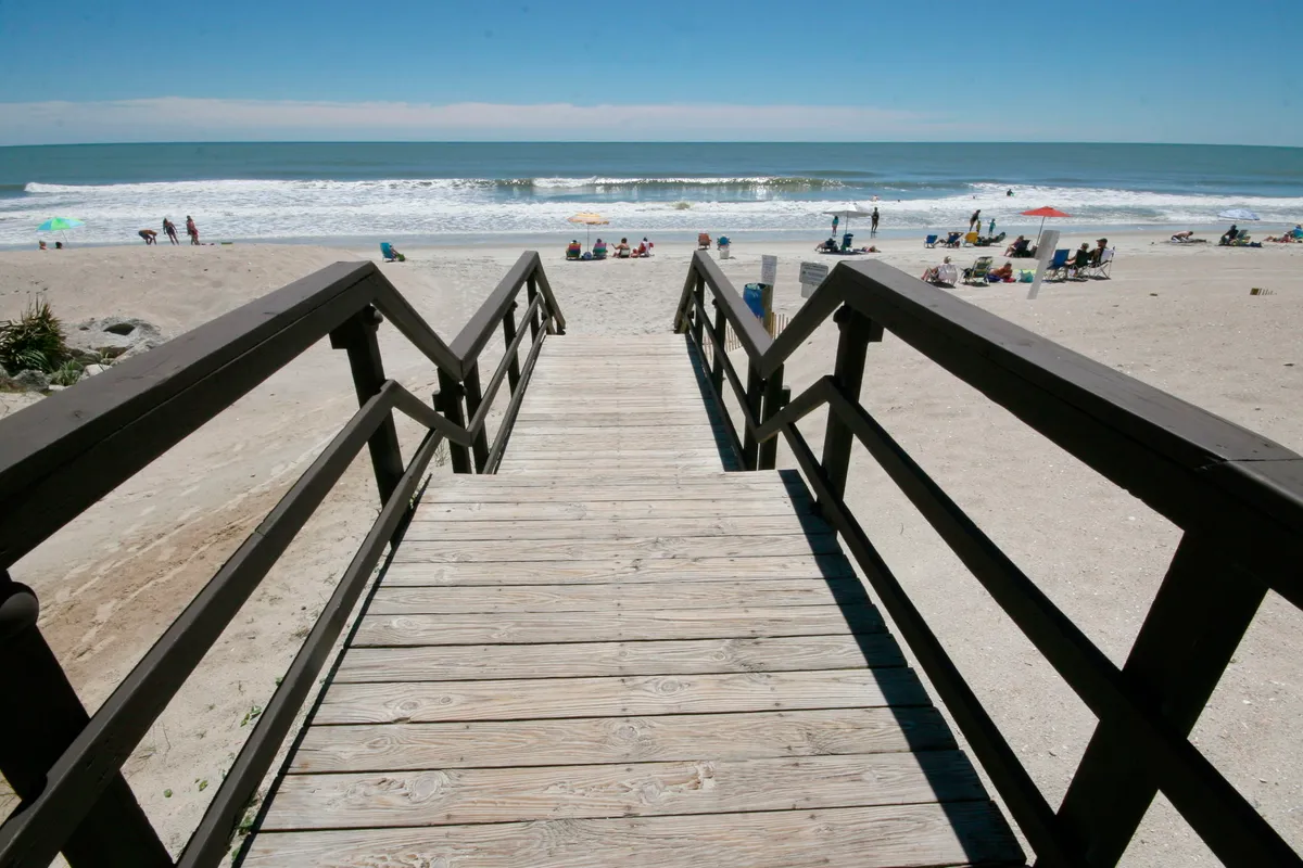 A scenic view of a wooden staircase leading down to a wide, sandy beach with gentle ocean waves, colorful umbrellas, and beachgoers enjoying the sun. The atmosphere is inviting and carefree, perfect for sandcastle building and wave jumping.