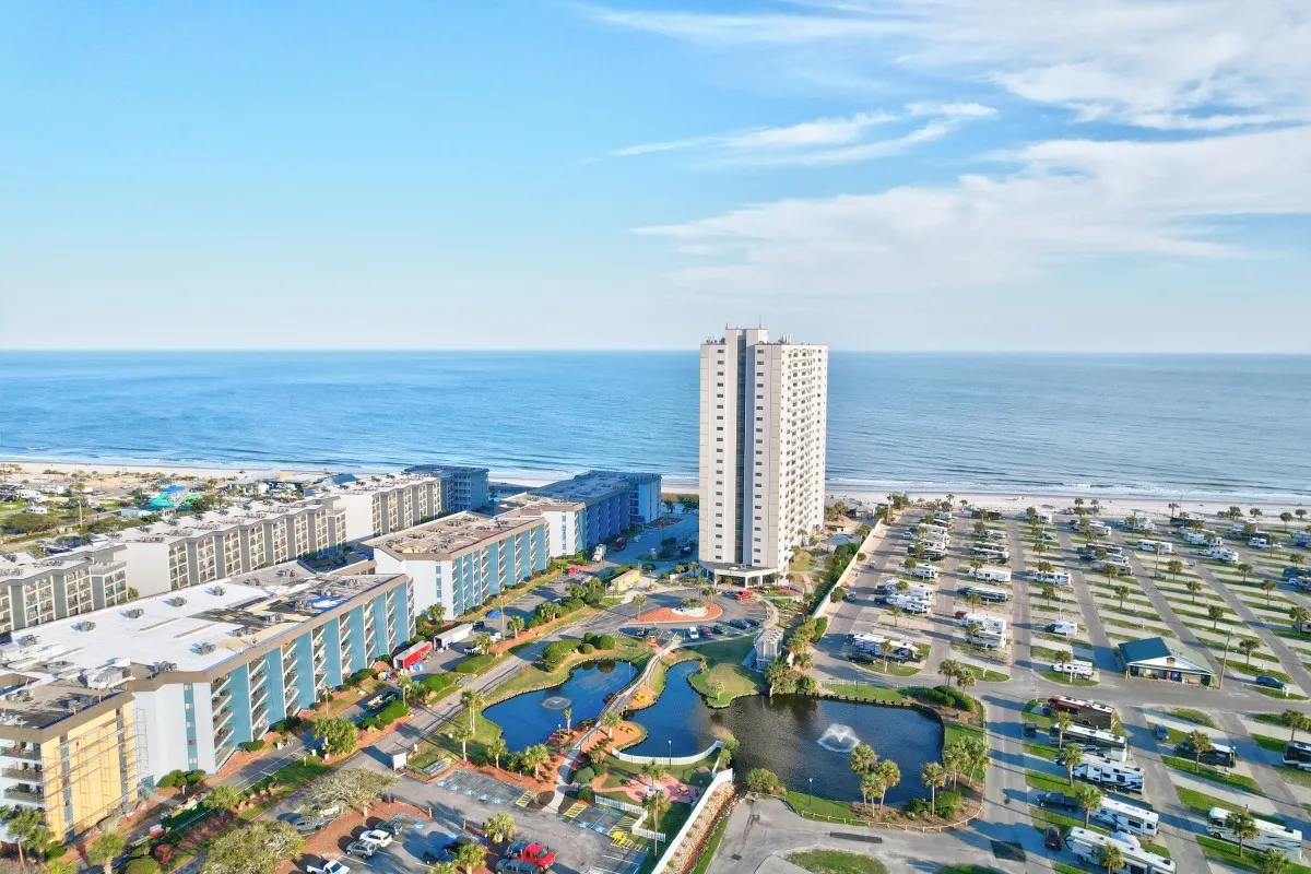 The photo features a tall residential building overlooking a spacious pool area, a man-made pond with a fountain, lush landscaping, and distant beach and ocean views. The setting is open and welcoming, with areas for swimming, picnicking, and relaxation.