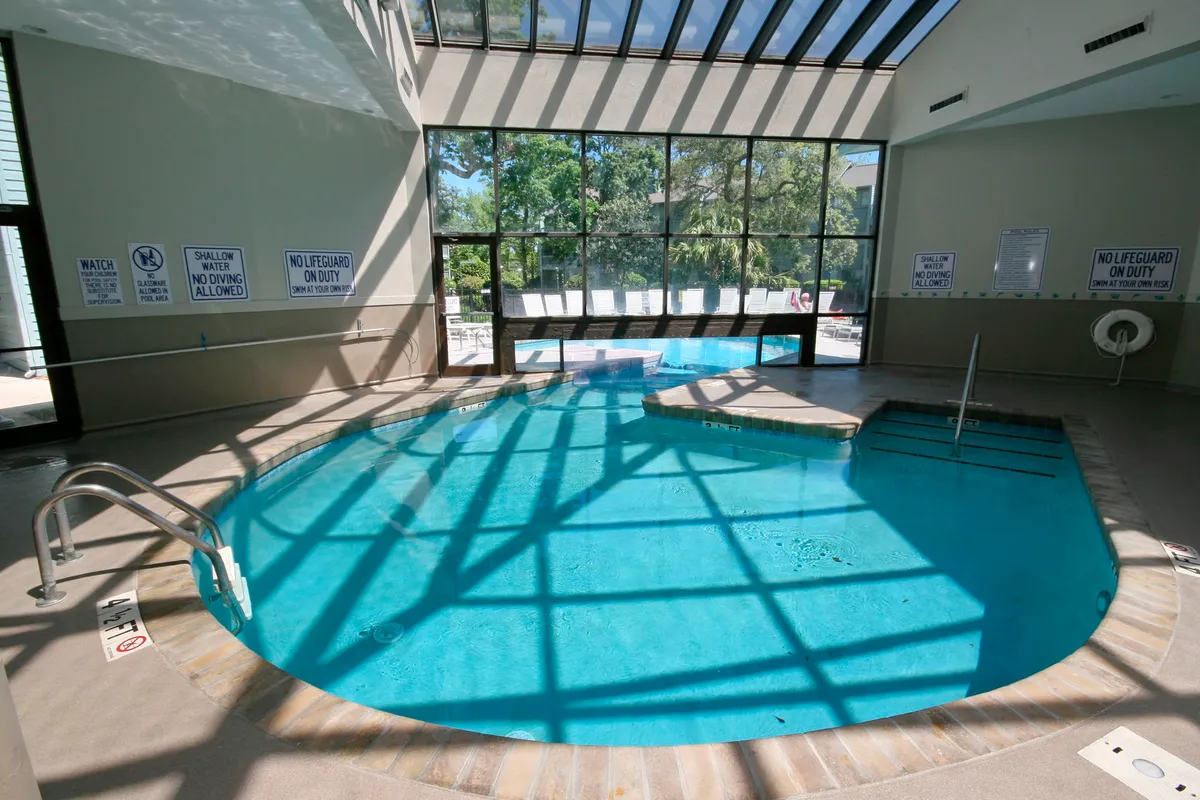 This image presents a bright and inviting indoor swimming pool with a glass roof, poolside seating, and clear signage for safety. The natural light streaming through the roof enhances a relaxing atmosphere, and the pool area looks clean and well-maintained.