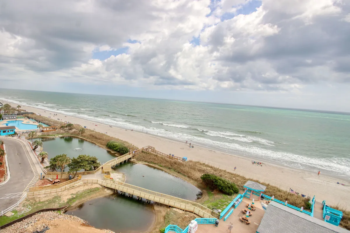 A wide-angle view from the property looking out over the ocean, sandy beach, and nearby boardwalk. Pools and people engaging in leisure activities can be seen in the foreground, with the serene expanse of the sea stretching into the distance. The overall mood is peaceful and enjoyable, evoking a sense of relaxation and possibility.