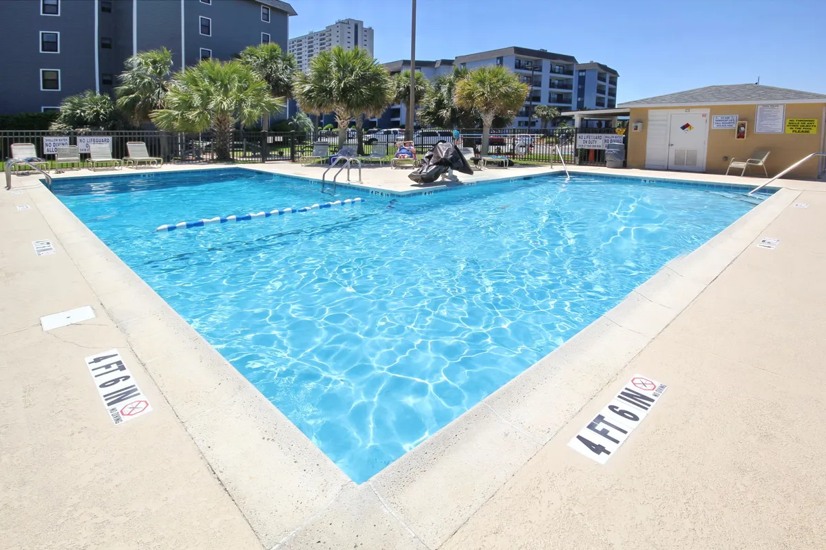 A sparkling outdoor swimming pool surrounded by palm trees and inviting poolside seating. A lifeguard chair is visible, and the atmosphere is cheerful with the suggestion of sunset lighting, evoking relaxation and romance.