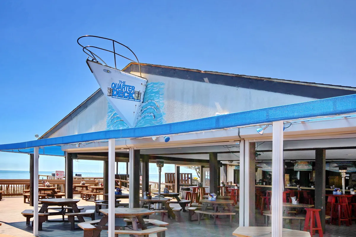 An exterior view of a seaside restaurant with outdoor seating and a scenic ocean backdrop. The scene captures a relaxed, cheerful mood, showing tables ready for guests to enjoy fresh seafood and the sound of the surf.