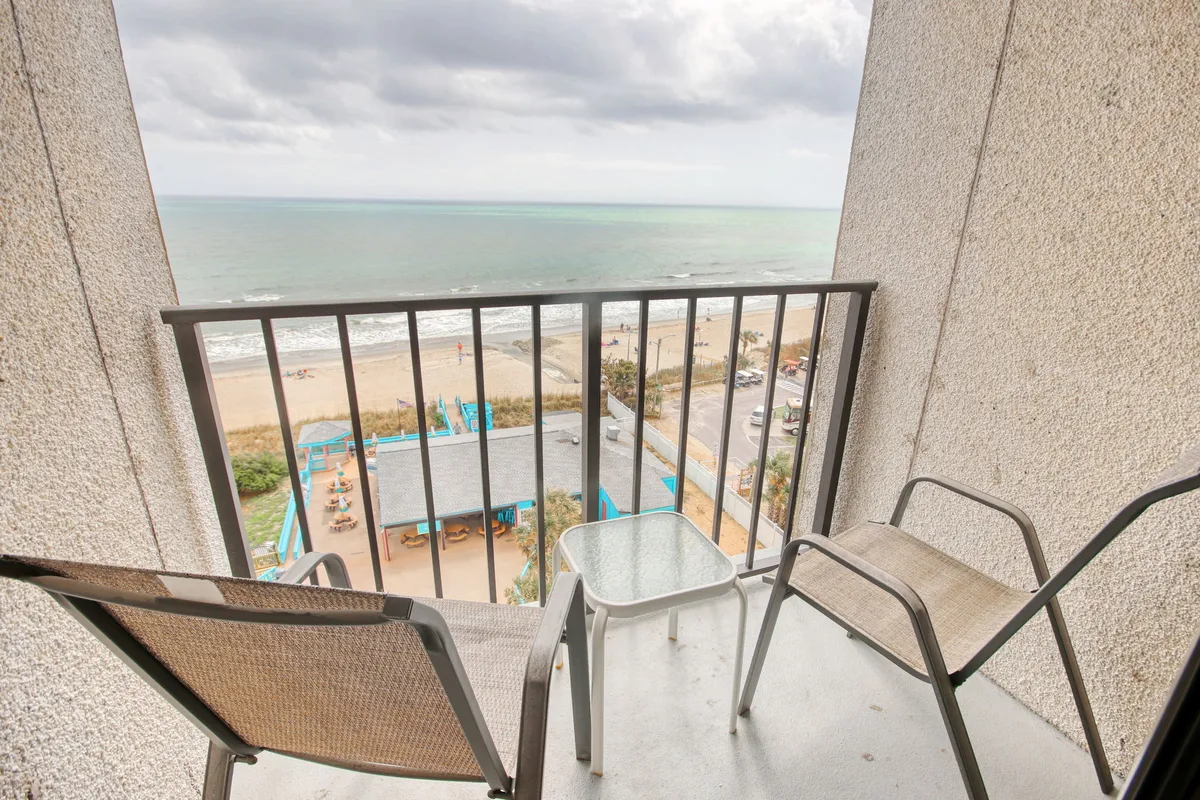 A serene private balcony setup with comfortable chairs and a small table, all overlooking an expansive ocean view. Gentle sunlight illuminates the space, and the open railing offers an uninterrupted vista of the water. The mood is calm and inviting, suggesting a perfect spot for morning coffee or evening relaxation.