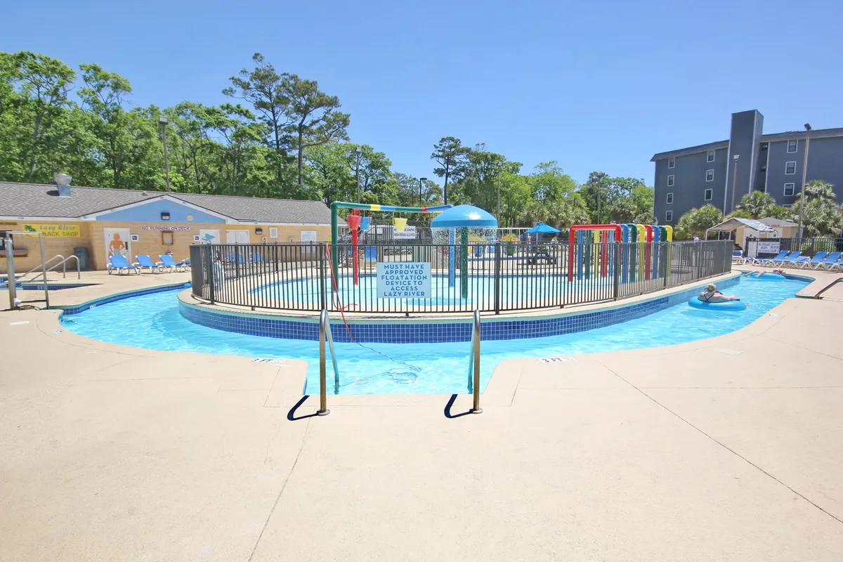 A vibrant outdoor pool scene featuring a large swimming pool, colorful water slides, surrounding trees, and plenty of lounge chairs. Children are seen playing and families are gathered around, enjoying a sunny day together.