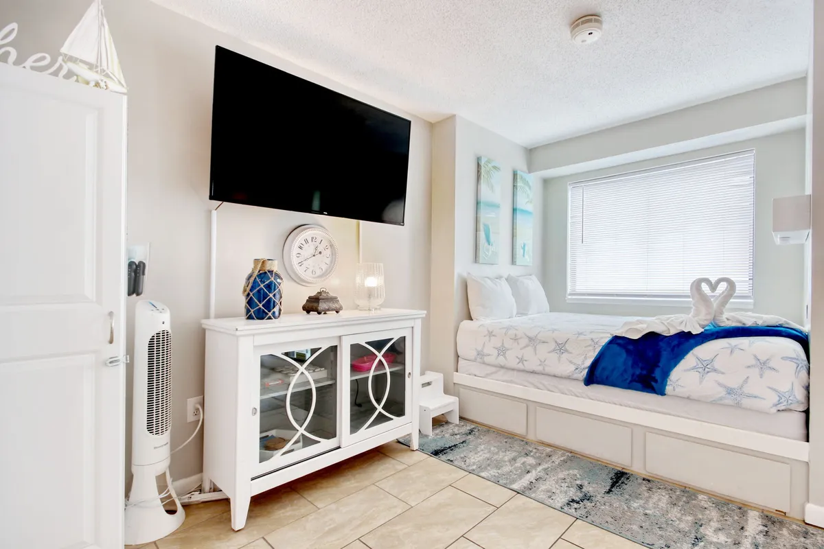 The photo shows a comfortable bed with a blue blanket, a television mounted on the wall, tasteful beach-themed artwork, and white furniture with glass doors. The aesthetic is serene and airy, with morning light filtering into the room.
