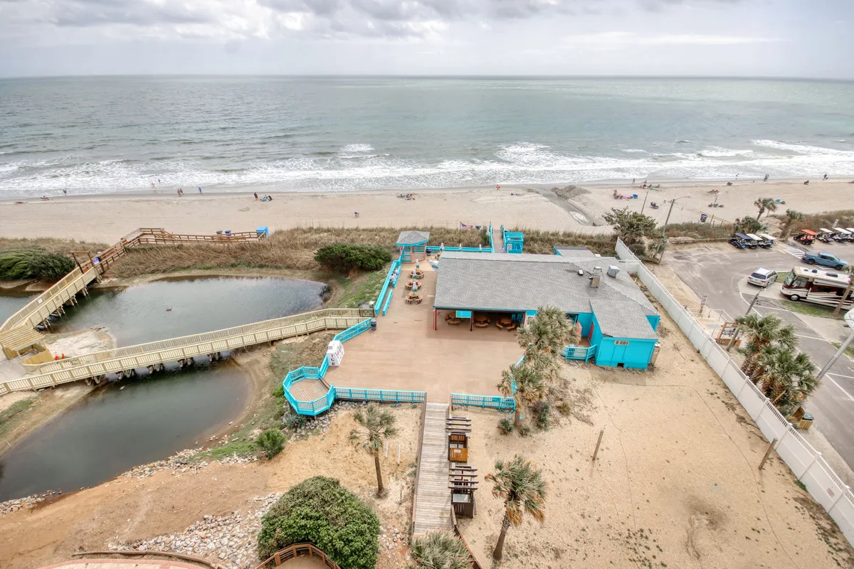 A colorful exterior shot featuring the beach, ocean, boardwalk, vibrant buildings, and lush vegetation. The scene captures the lively yet laid-back atmosphere of Myrtle Beach and hints at the diverse activities available just steps from the property.