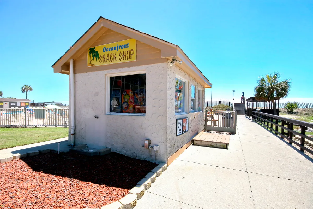 A cheerful exterior shot of an oceanfront snack shop with vibrant window displays, surrounded by palm trees and a pathway leading to the beach. The scene suggests easy access to refreshments and social spaces just steps from the sand.