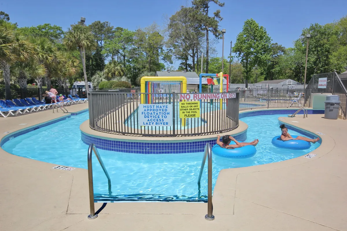 A vibrant exterior pool area featuring a winding lazy river, swimming pool, play space, loungers, and clear signage. The scene captures guests floating, sunbathing, and enjoying various water activities.
