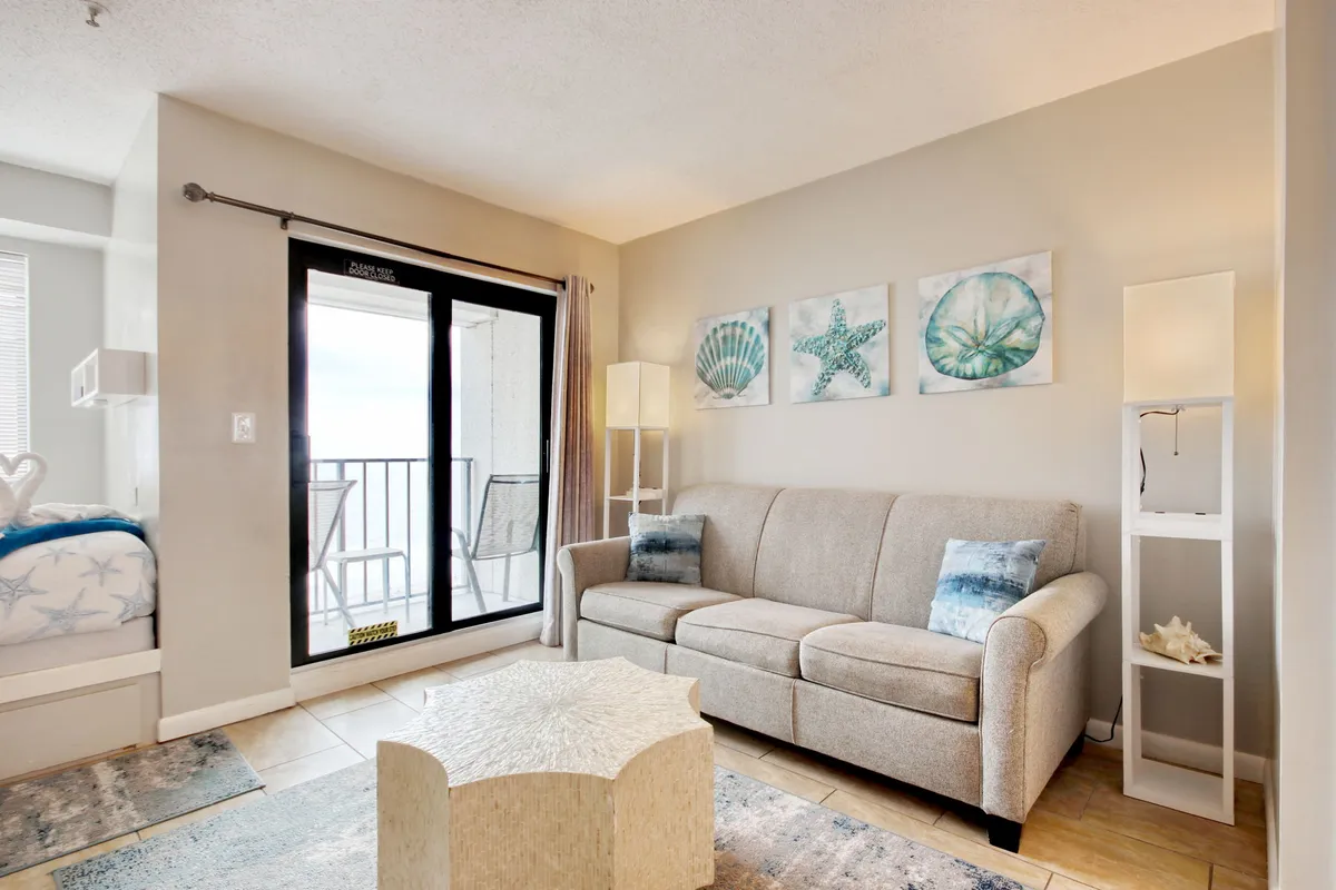 The image showcases a sunlit studio living area with a cozy sofa, ocean-themed artwork, and a coffee table arranged before a sliding glass door. The door opens to a private balcony overlooking the ocean, with soft, natural light filling the space and creating a tranquil, inviting atmosphere.